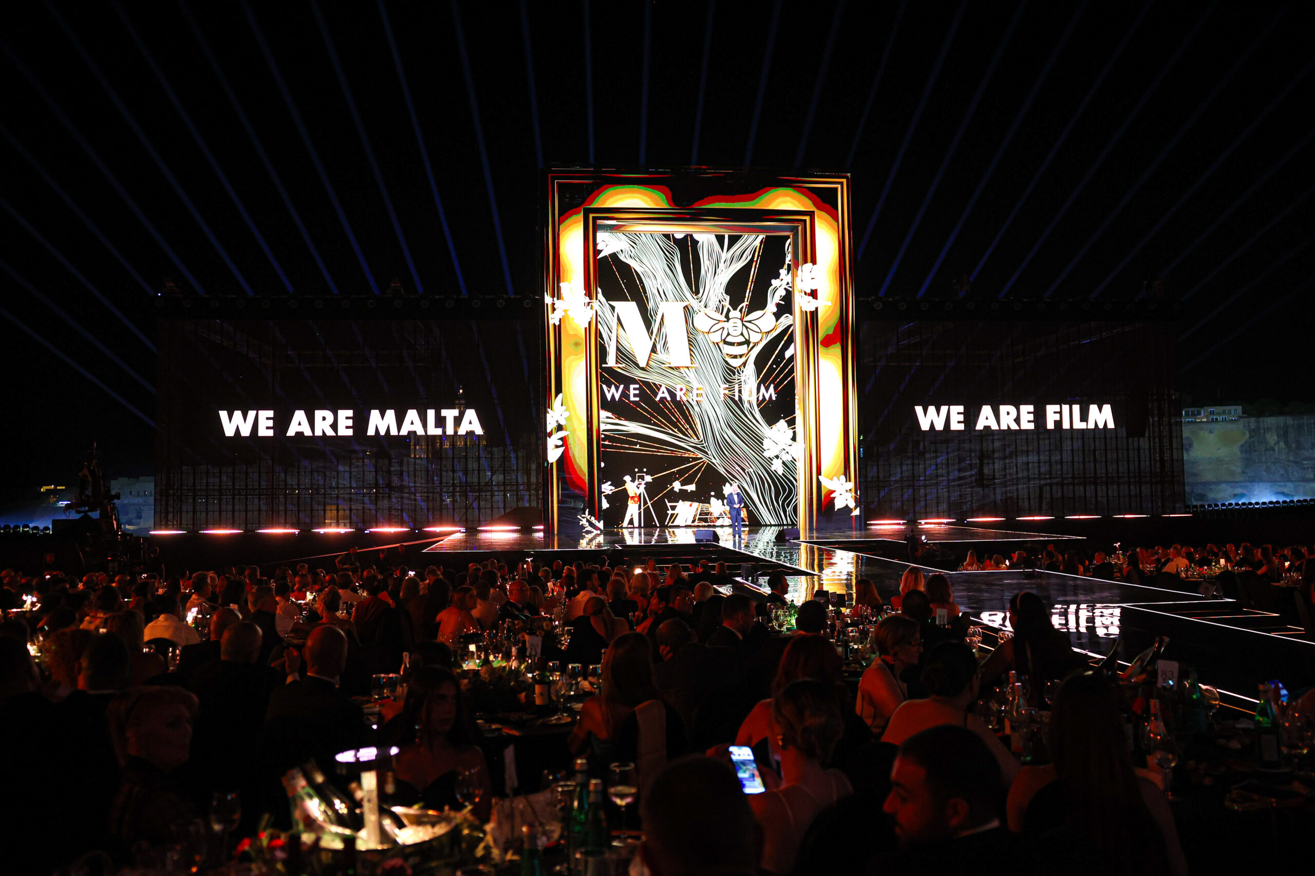 The stage at Mediterranean Film Festival is lit up in a dark room for the awards ceremony. The left side says We Are Malta and the right says We Are Film.