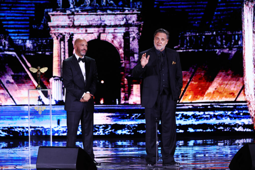 Russell Crowe gives a speech onstage at the Golden Bee Awards Ceremony at MFF, Maltese Film Commissioner Johann Grech stands before him to the left