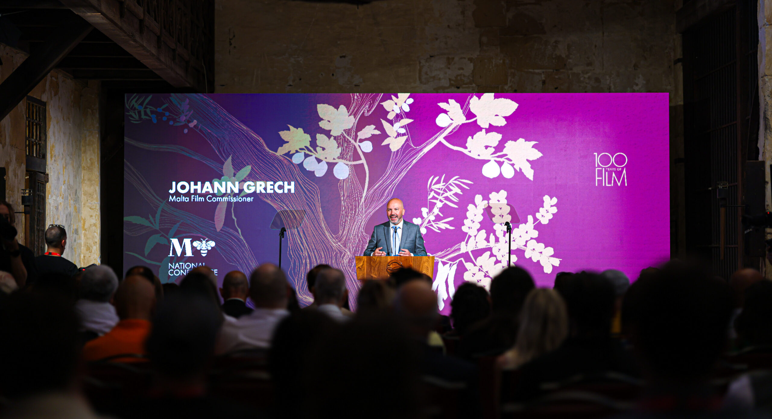 Johann Grech, Malta Film Commissoner, stands onstage in front of a screen in front of an audience