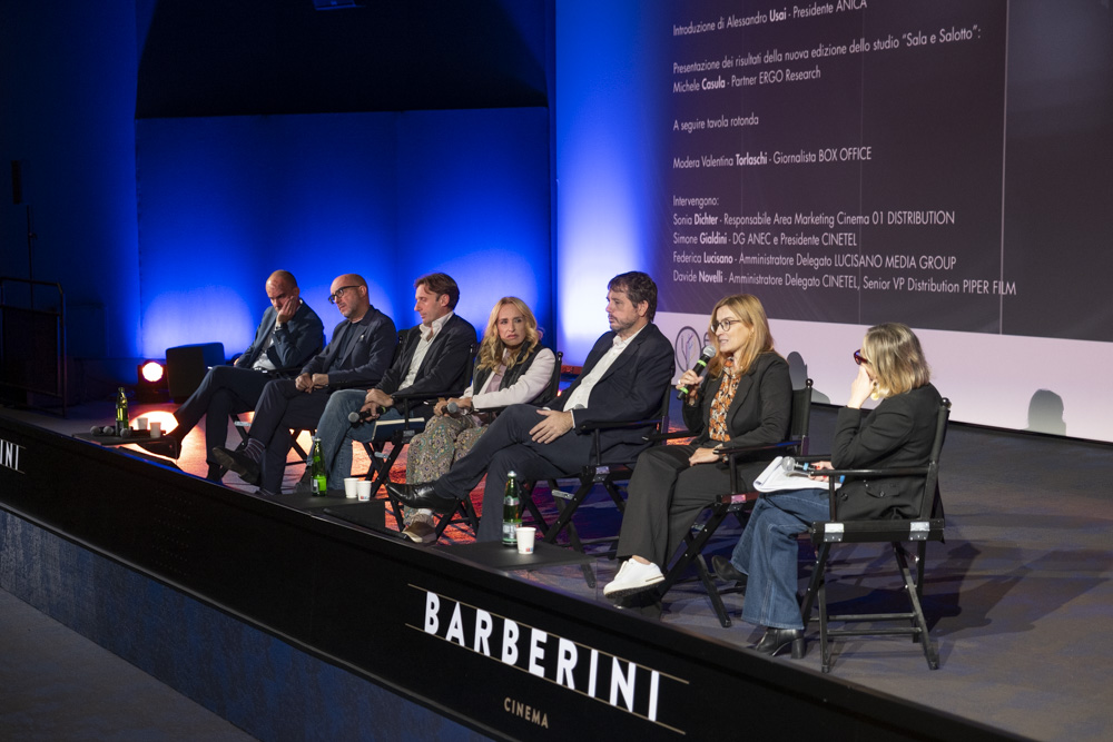 The panellists sit on stage with a screen behind them, from left to right: Michele Casula; Alessandro Usai; Davide Novelli; Federica Lucisano; Simone Gialdini; Sonia Dichter; Valentina Torlaschi