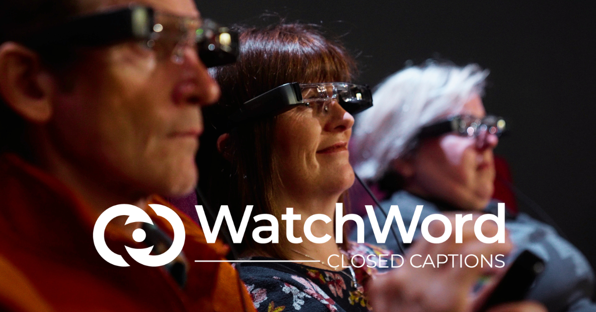 Three audience members sit looking at the big screen, while wearing WatchWord smart glasses. The "WatchWord Closed Captions” logo in white sits at the bottom of the image, under their faces.